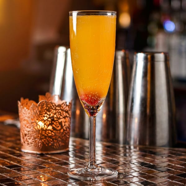 A vibrant orange cocktail with raspberries sits on a tiled bar next to a glowing candle.