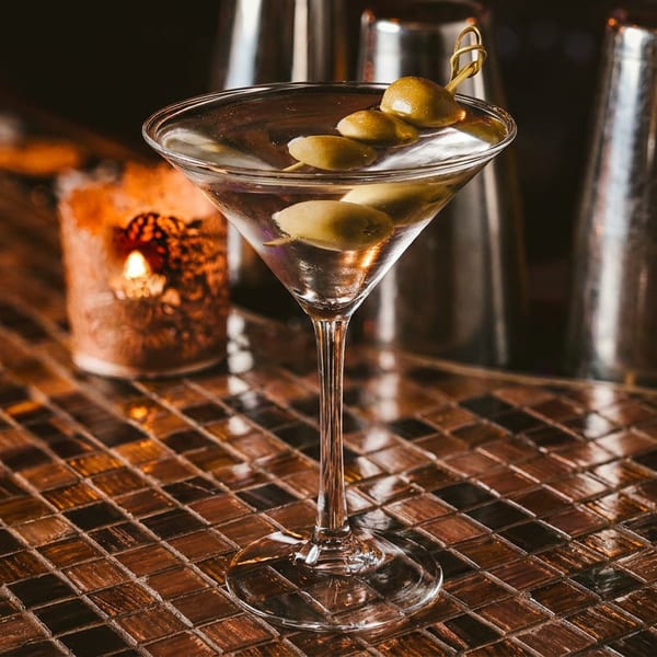 Martini with olives on a skewer, on a tiled bar next to a lit candle and shakers.
