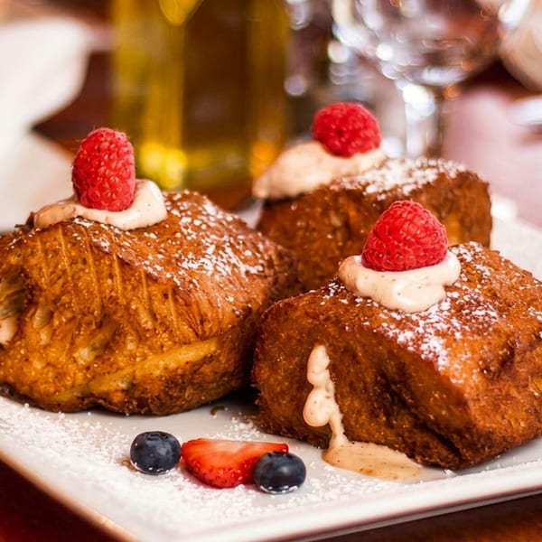 Three golden-brown French toast pieces topped with whipped cream, raspberries, and powdered sugar, with berries.