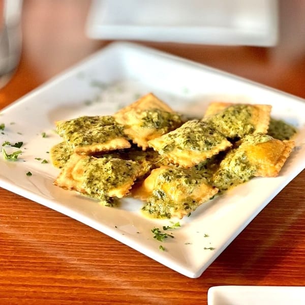 Square plate of golden ravioli topped with green pesto sauce and parsley on a wooden table.