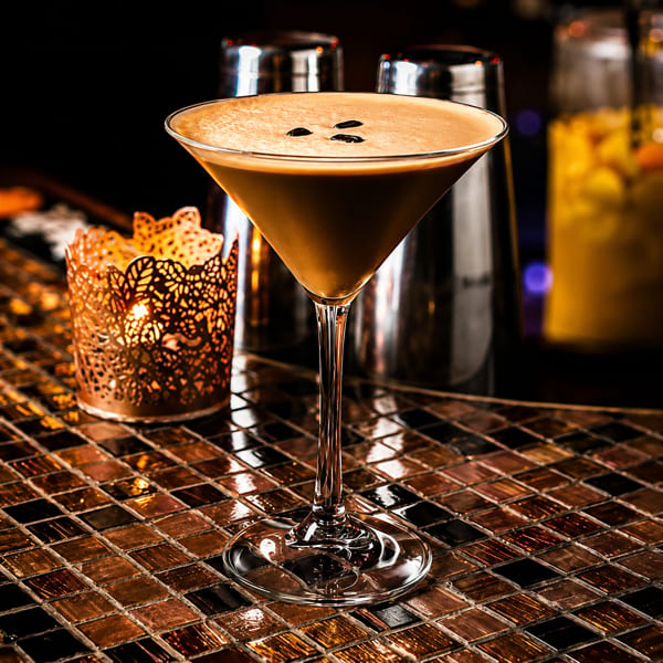 Espresso martini with coffee beans on a tiled bar next to a glowing candle holder.