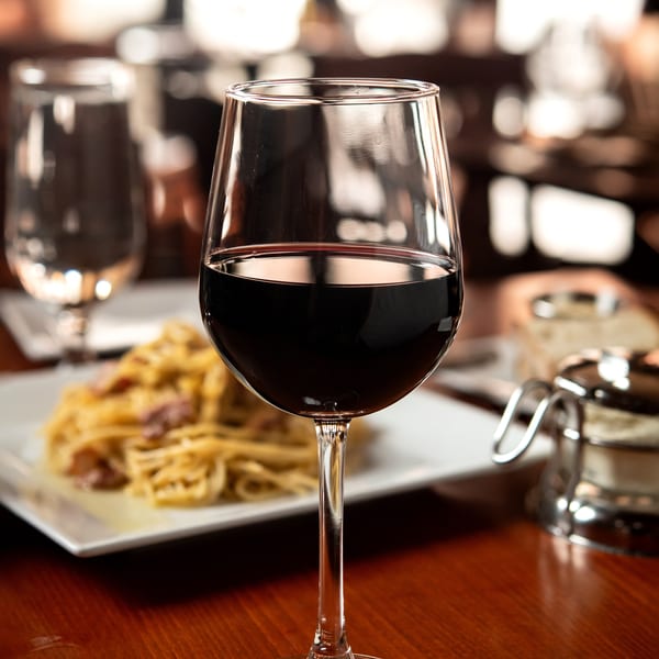 A glass of red wine on a wooden table, with a plate of pasta in the background.