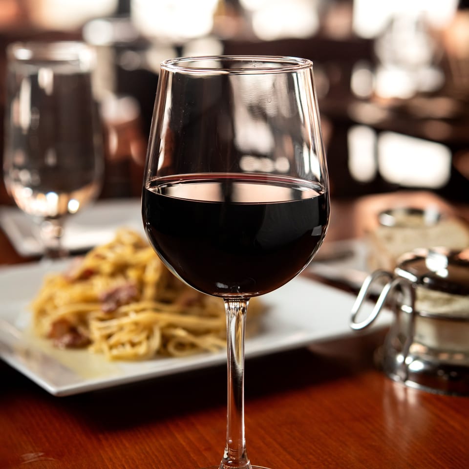 A glass of red wine on a wooden table, with a plate of pasta in the background.