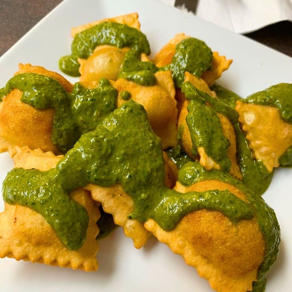 Golden-brown fried ravioli generously topped with vibrant green pesto sauce on a white plate.
