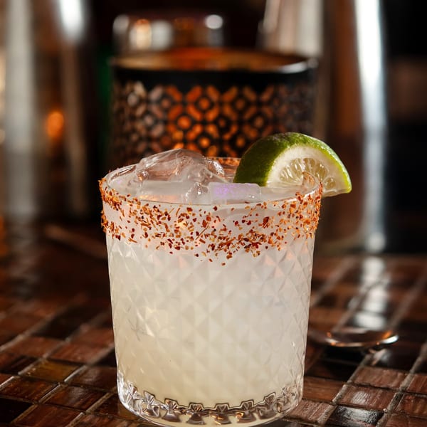 A Mama Mia 44SW cocktail with a chili-salt rim, ice, and a lime wedge on a dark bar.