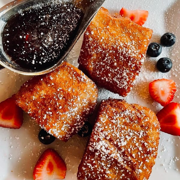 Three powdered sugar-dusted Italian fritters with strawberries, blueberries, and a side of syrup.