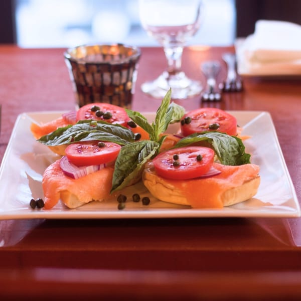 Four bruschetta with smoked salmon, tomato, red onion, capers, and basil on a white plate.